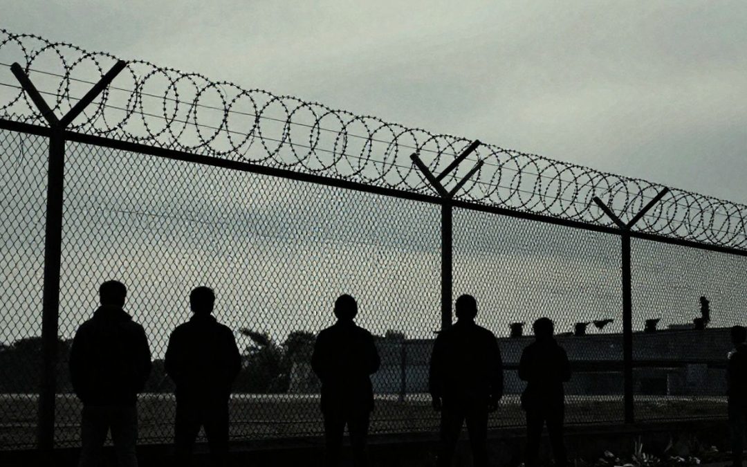 Detention center fence with silhouetted figures behind it.