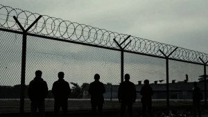 Detention center fence with silhouetted figures behind it.