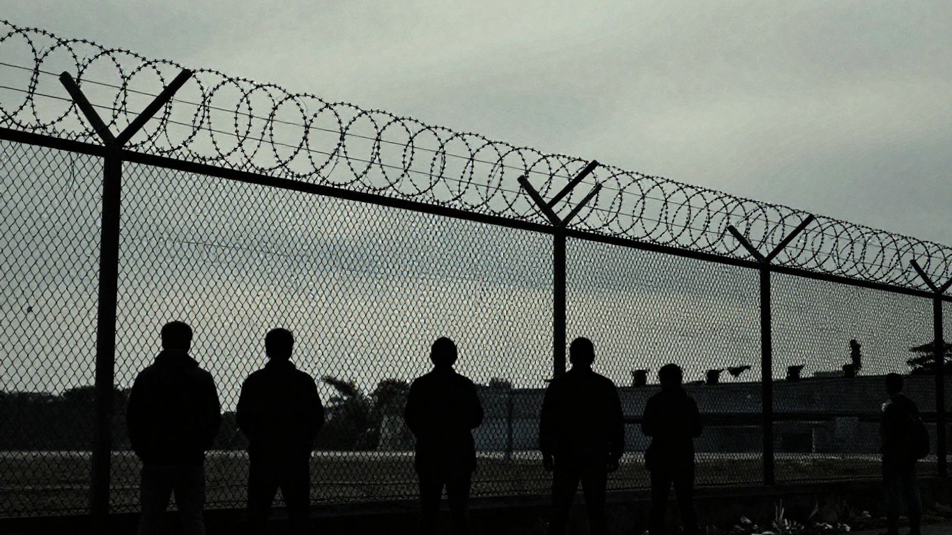 Detention center fence with silhouetted figures behind it.